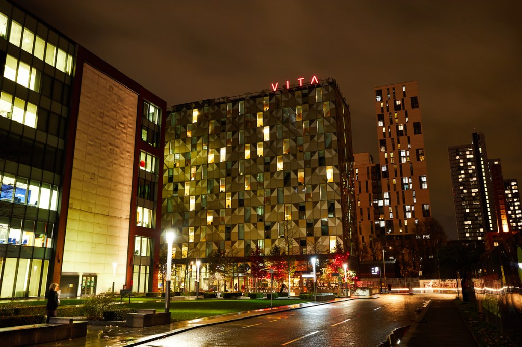 A modern city street at night features illuminated buildings, including one with a geometric facade and a red "VITA" sign—popular accommodation for students moving to Manchester. Trees with autumn leaves and light trails from passing cars complete the scene.
