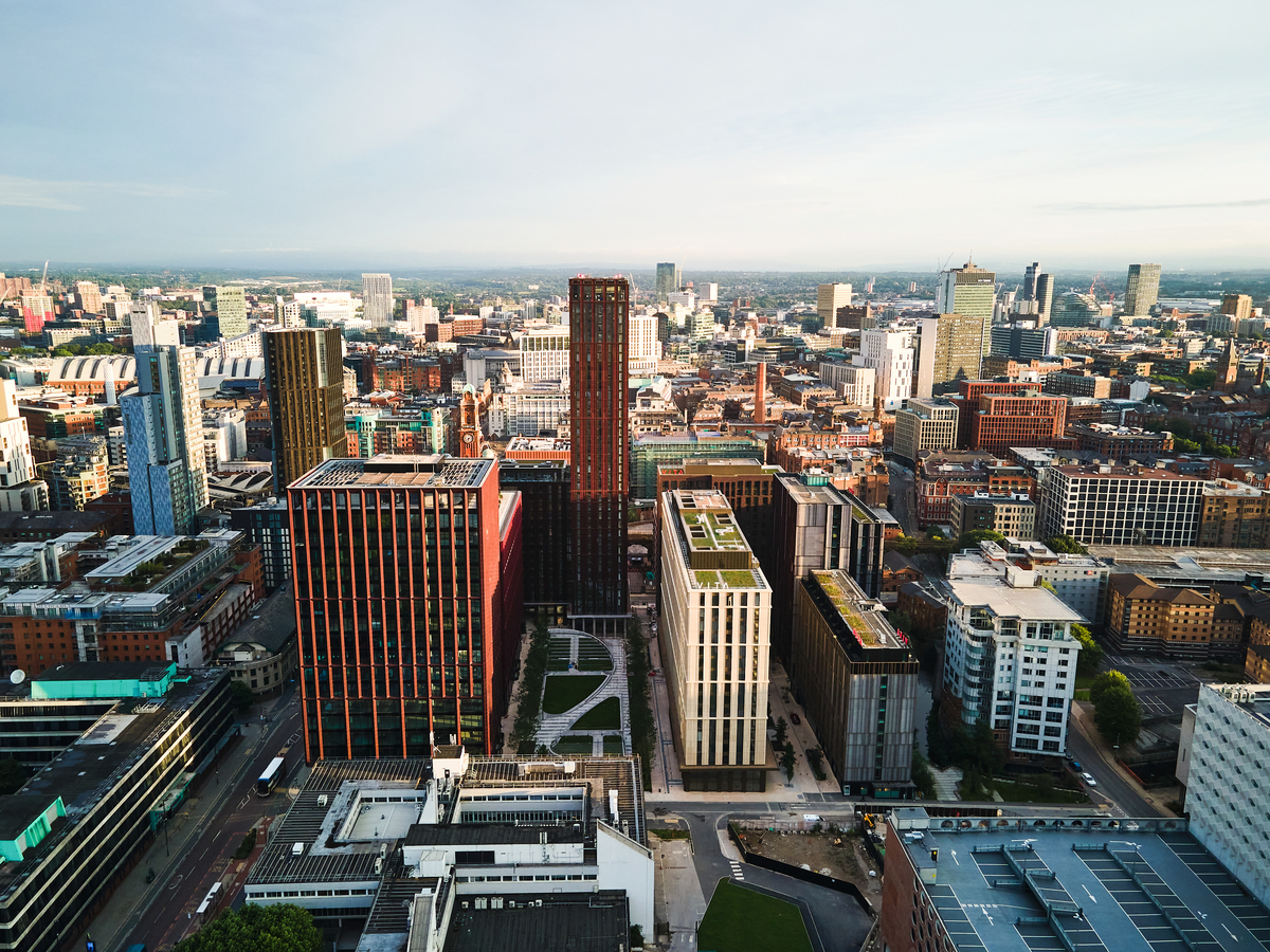 Aerial view of a modern city with tall office buildings, green rooftops, and urban streets—ideal for students moving to Manchester seeking accommodation—set against a wide skyline under a mostly clear sky.
