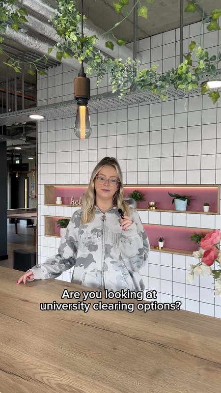 A woman wearing glasses and a camouflage hoodie stands in a modern, plant-decorated room, holding a pen. Text on the image reads, "Are you looking at university clearing options?.