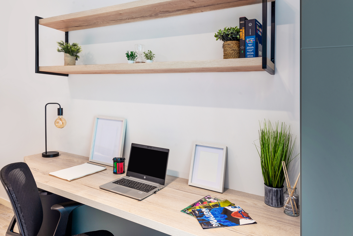 A modern, tidy desk setup with a laptop, notebook, chair, pen holder, and magazines. Above the desk is a wooden shelf with potted plants and books. The area is well-lit and minimalistic, creating a productive workspace.