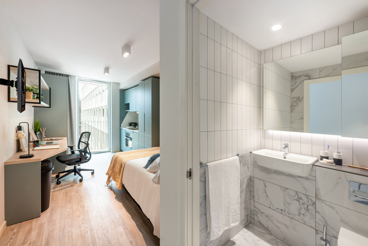 A modern, well-lit studio apartment with a workspace, bed, and kitchen area on the left, and a sleek bathroom with white tile, a sink, and a towel hanging on the right.