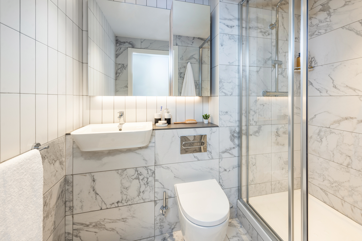 Modern bathroom with marble tile walls, a wall-mounted sink, a toilet, a glass-enclosed shower, and a large mirror above the sink. Accessories include soap dispensers and a small plant on the counter.