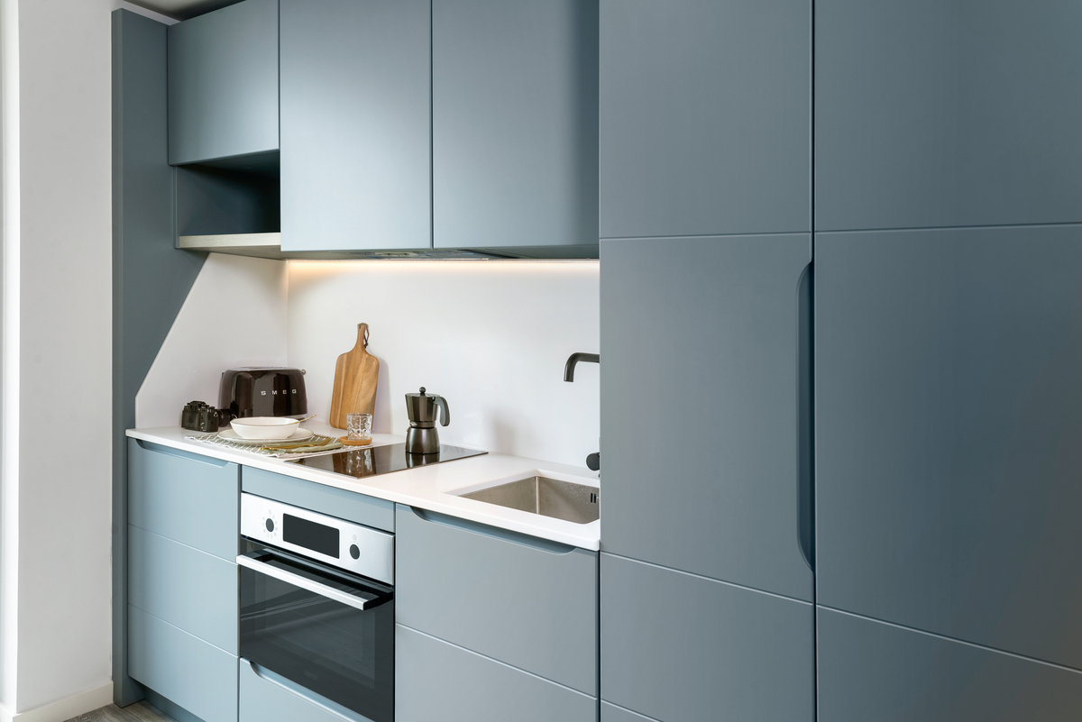 A modern kitchen with matte blue cabinets, a white countertop, a built-in oven, sink, toaster, coffee maker, and a wooden cutting board against the backsplash under soft under-cabinet lighting.