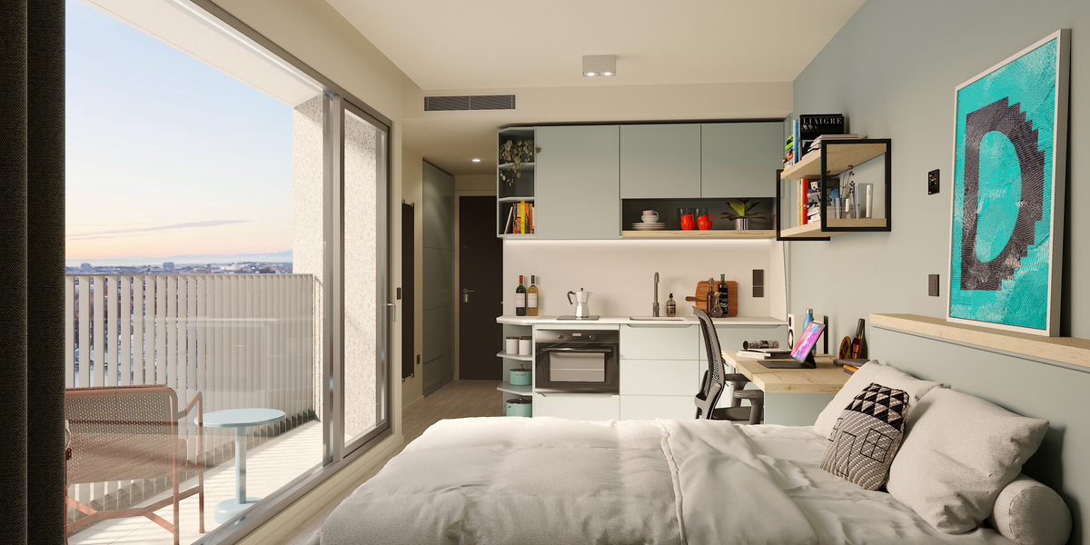 A modern studio apartment with a neatly made bed, kitchenette, desk, shelves, and a balcony with a city view at sunset. The room is decorated with art, books, and plants, and has light, neutral colors.