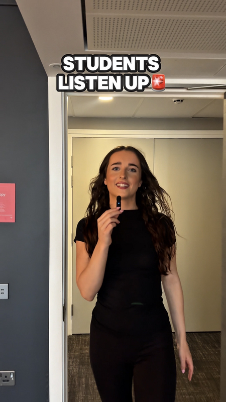 A person with long brown hair, dressed in black, stands indoors holding a marker and smiling. Text above reads, “STUDENTS LISTEN UP” with a red alert emoji.