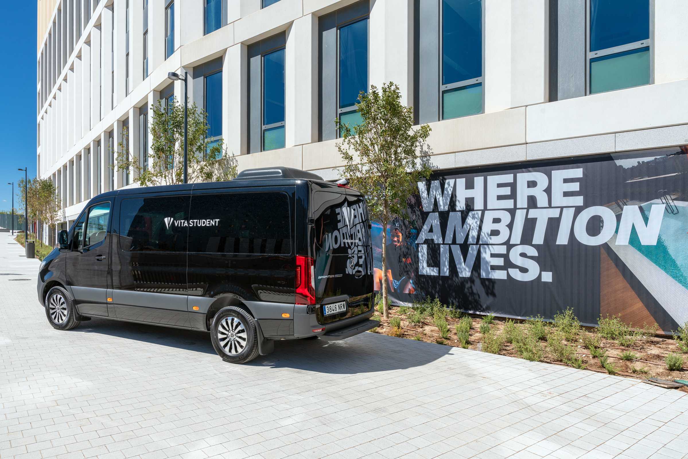 A black Vita Student van is parked beside a modern building. A large banner on the building reads “WHERE AMBITION LIVES.” Some small trees are planted along the walkway.