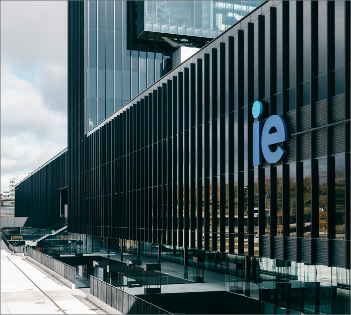 A modern black glass building with vertical slats, featuring a large blue "ie" logo, stands in an urban setting. The sky is partly cloudy. This stylish spot is ideal for those seeking student accommodation in Madrid.