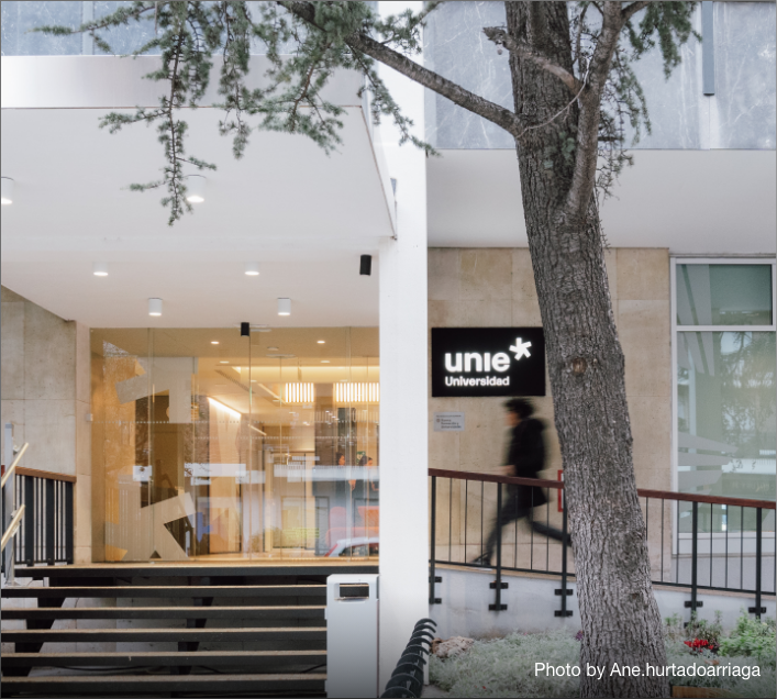 A person walks past the entrance of a modern building with a sign that reads "unie Universidad." Glass doors and steps lead up to the entrance, with Student Accommodation in Madrid located nearby and a tree standing in the foreground.