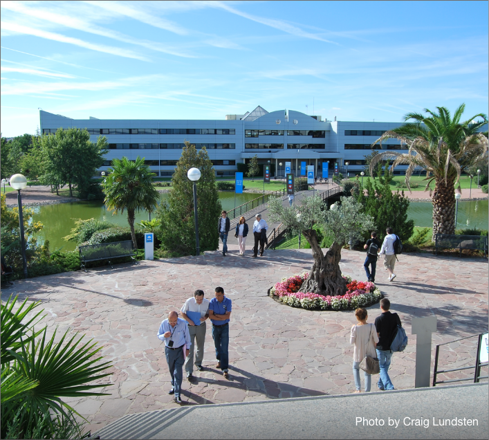 People walk on a stone path lined with palm trees and greenery, heading toward a modern office building across a small lake—evoking the feel of premium student accommodation in Madrid. "Photo by Craig Lundsten" appears in the lower right corner.