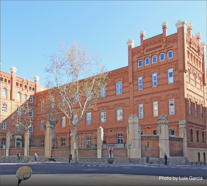A large, historic red-brick building with tall windows and decorative battlements, this impressive Student Accommodation in Madrid sits behind a fence and trees on a sunny day, with pedestrians and parked cars visible out front.