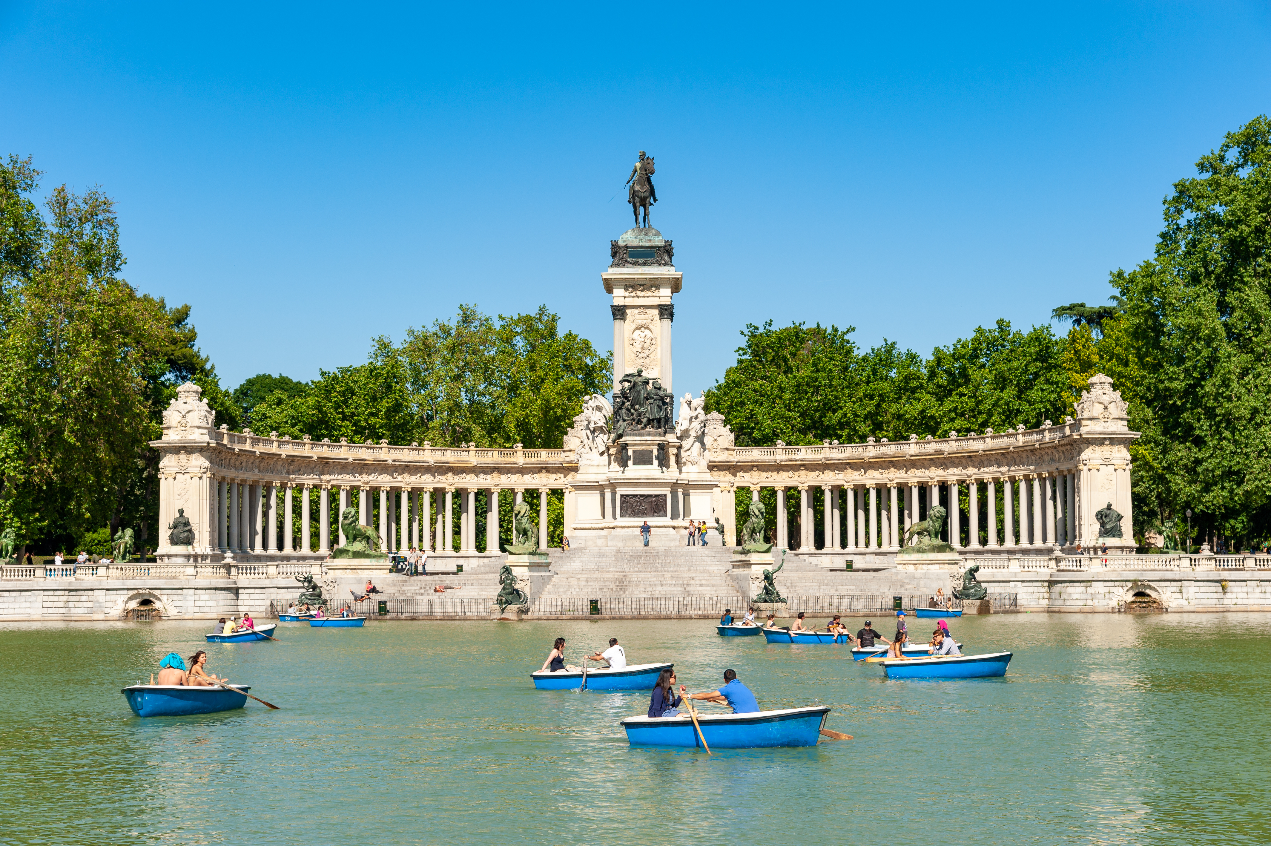 People row blue boats on a large pond in front of the semicircular Monument to Alfonso XII in Madrid’s Retiro Park, surrounded by green trees under a clear blue sky.
