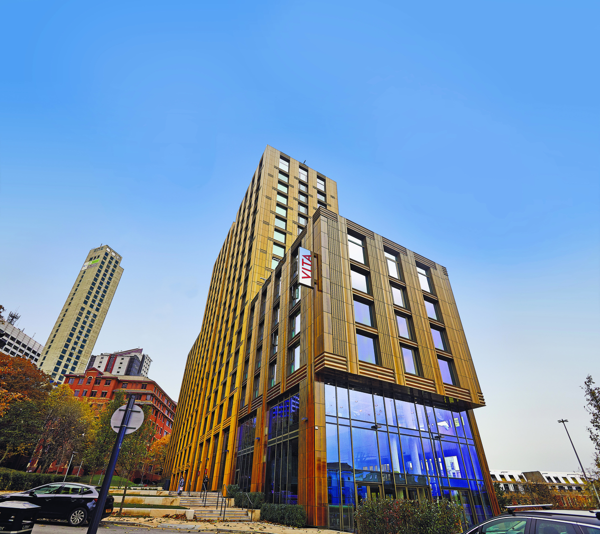 A modern, tall building with a "Vita" sign on the corner, featuring large glass windows on the lower floors, stands against a clear blue sky—an ideal accommodation choice for students awaiting university admission through UCAS.