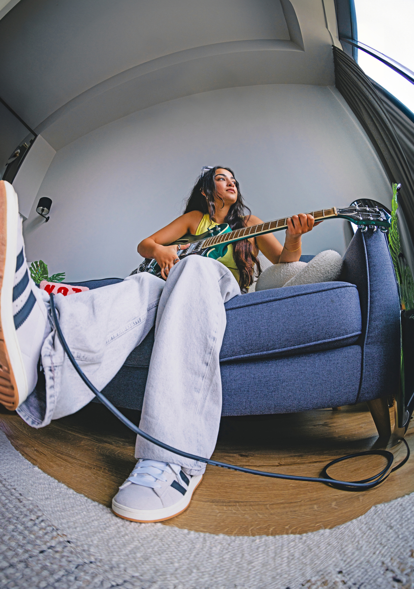 A young woman sits on a blue sofa playing an electric guitar, her foot resting on the floor as a guitar cable curves in the foreground—a quiet moment indoors, possibly reflecting between stages of the university admissions process.