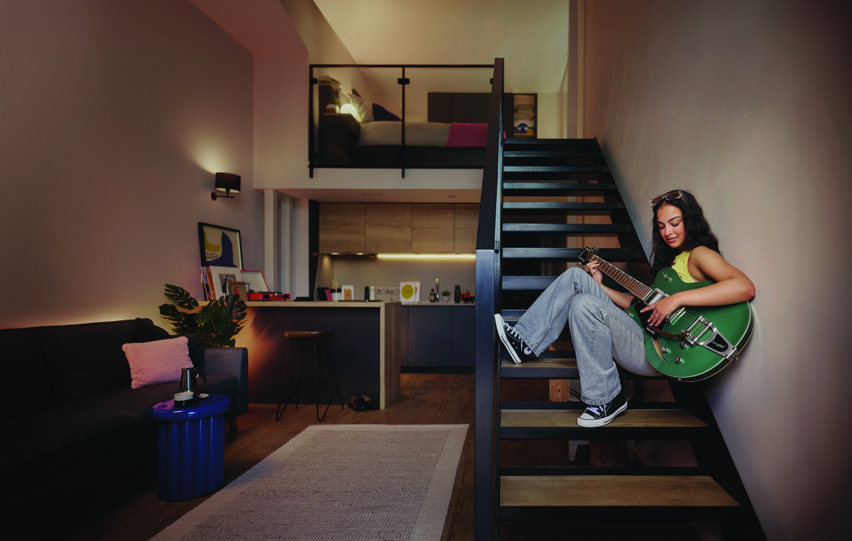 A young woman sits on stairs in a modern apartment, playing a green electric guitar. The apartment features a lofted bed, a kitchen, a sofa, and contemporary decor. Natural light brightens the cozy space.