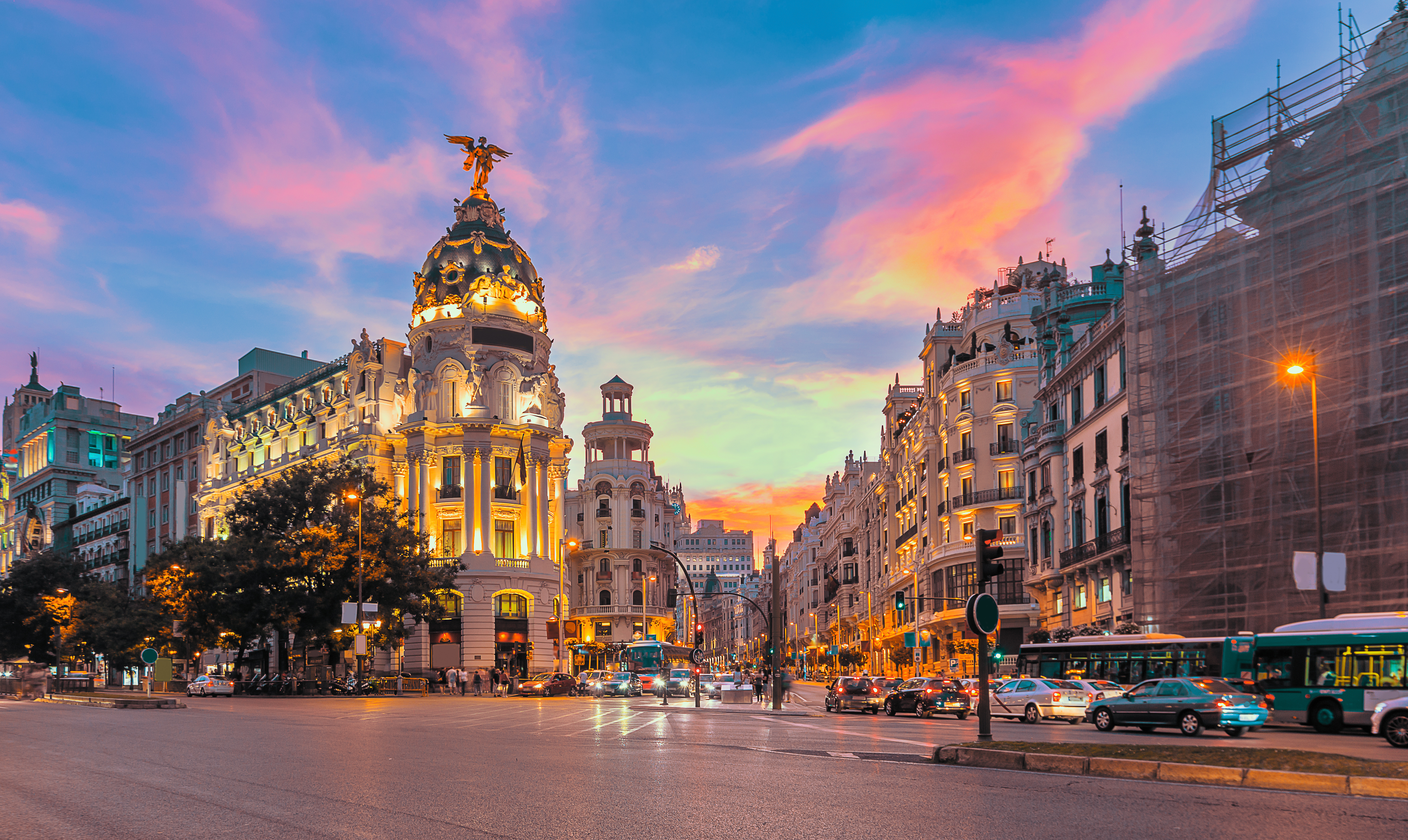The Gran Via in Madrid at sunset, featuring the illuminated Metropolis Building with its ornate dome and statue, surrounded by historic buildings, cars, and vibrant, colorful skies.