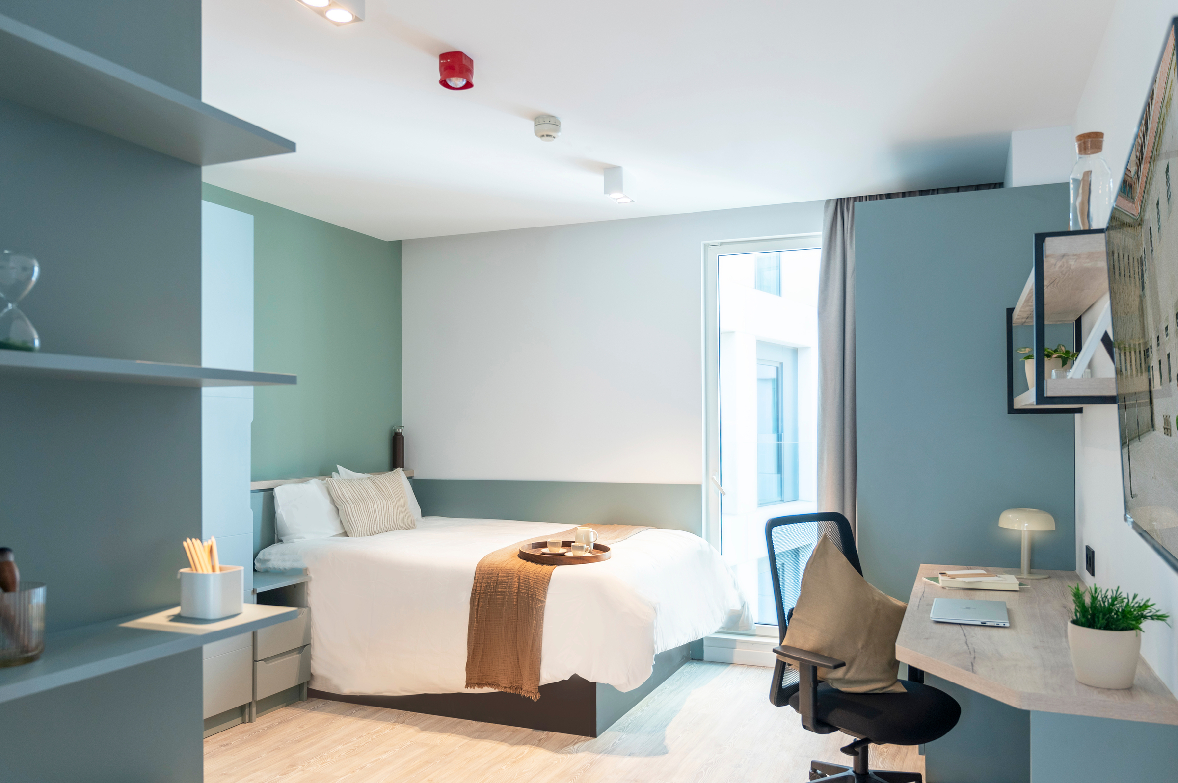 Modern, tidy bedroom with a neatly made bed, a tan throw, desk with chair, shelves, lamp, and large window letting in natural light. Decor is minimalist with soft green and neutral tones.