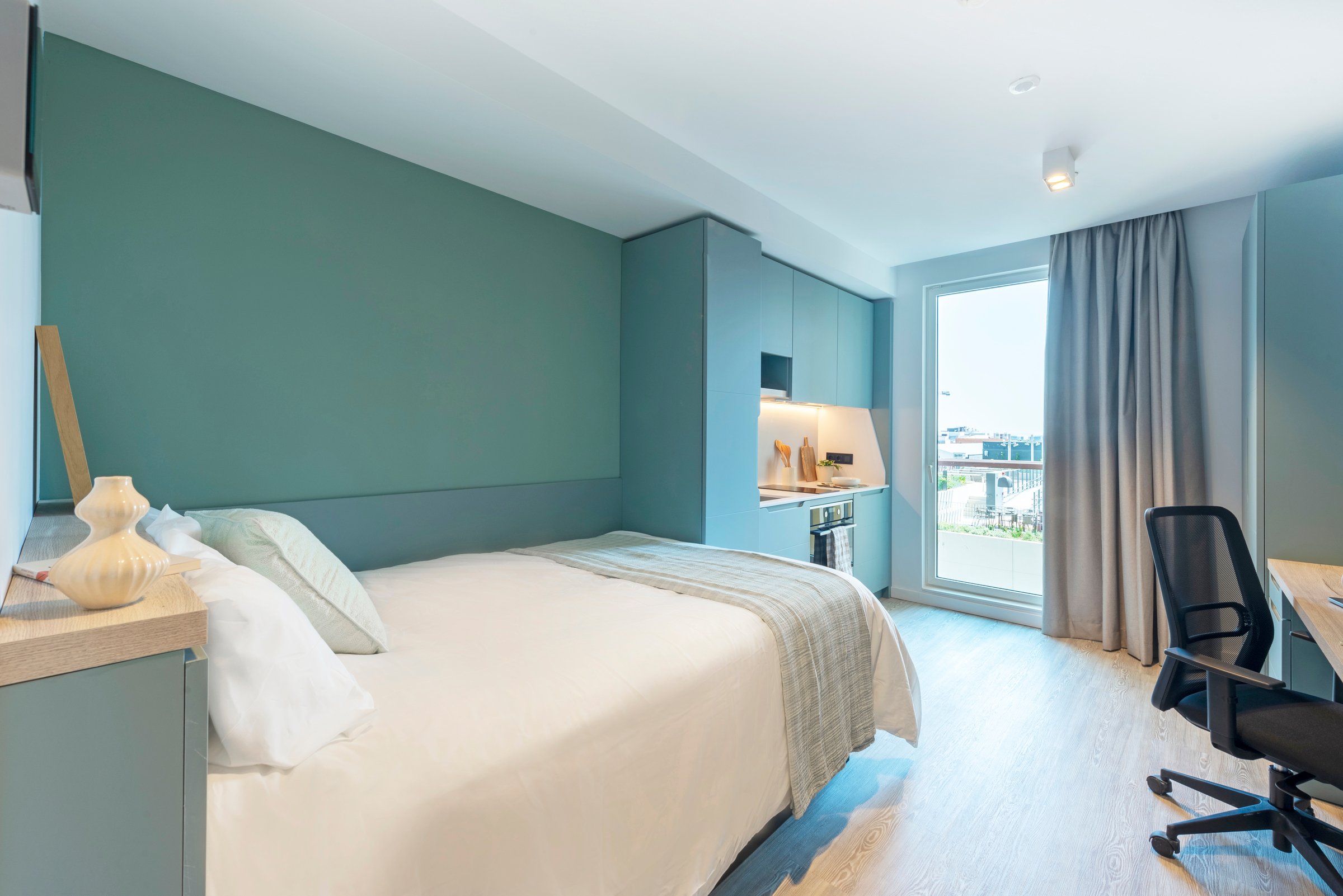 A modern bedroom with a neatly made bed, blue-green walls, a desk and chair, shelves, and a large window with curtains letting in natural light.