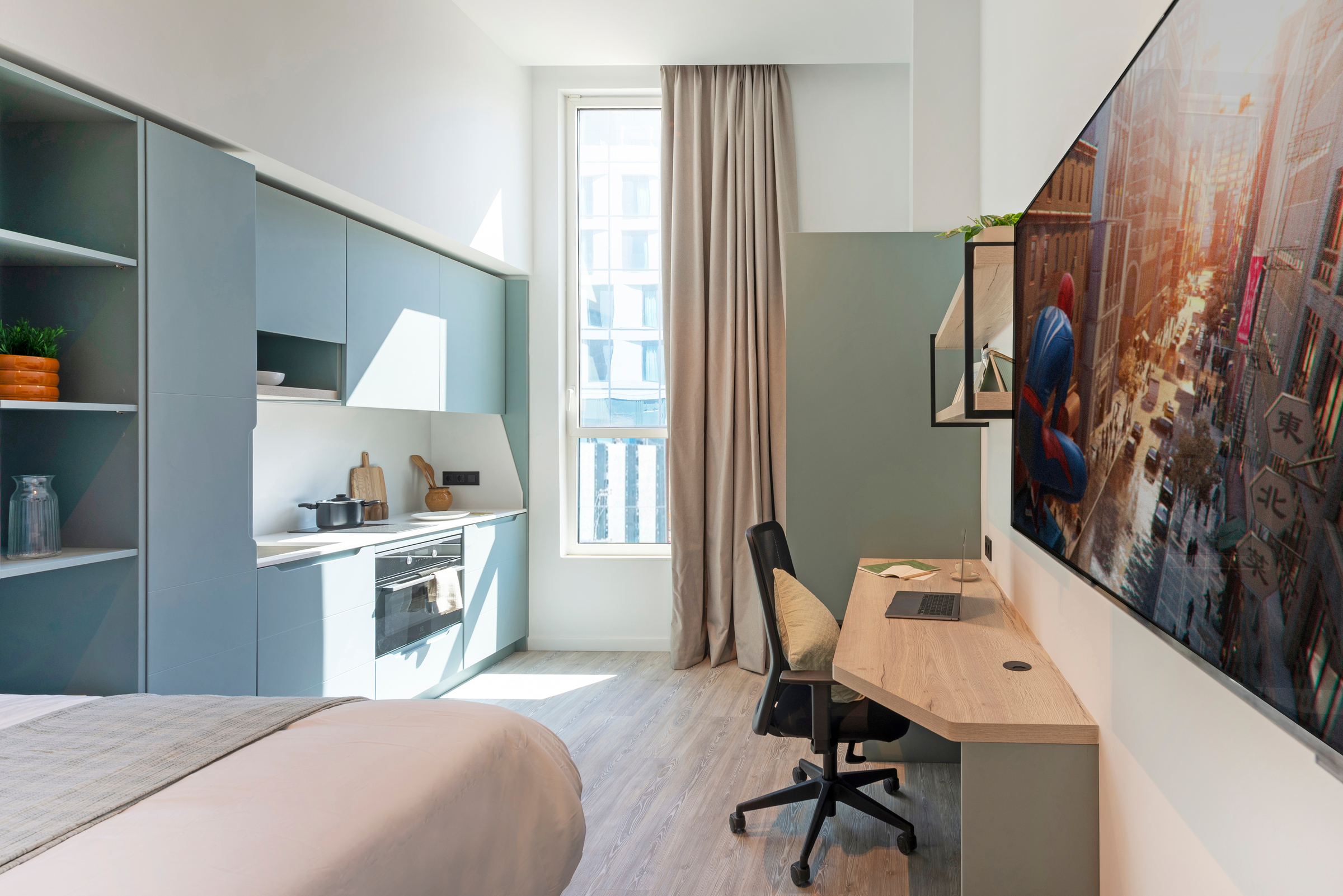 Modern studio apartment with a bed, built-in kitchen, desk, office chair, large wall-mounted TV displaying a Spider-Man scene, and a tall window with sheer curtains letting in natural light.
