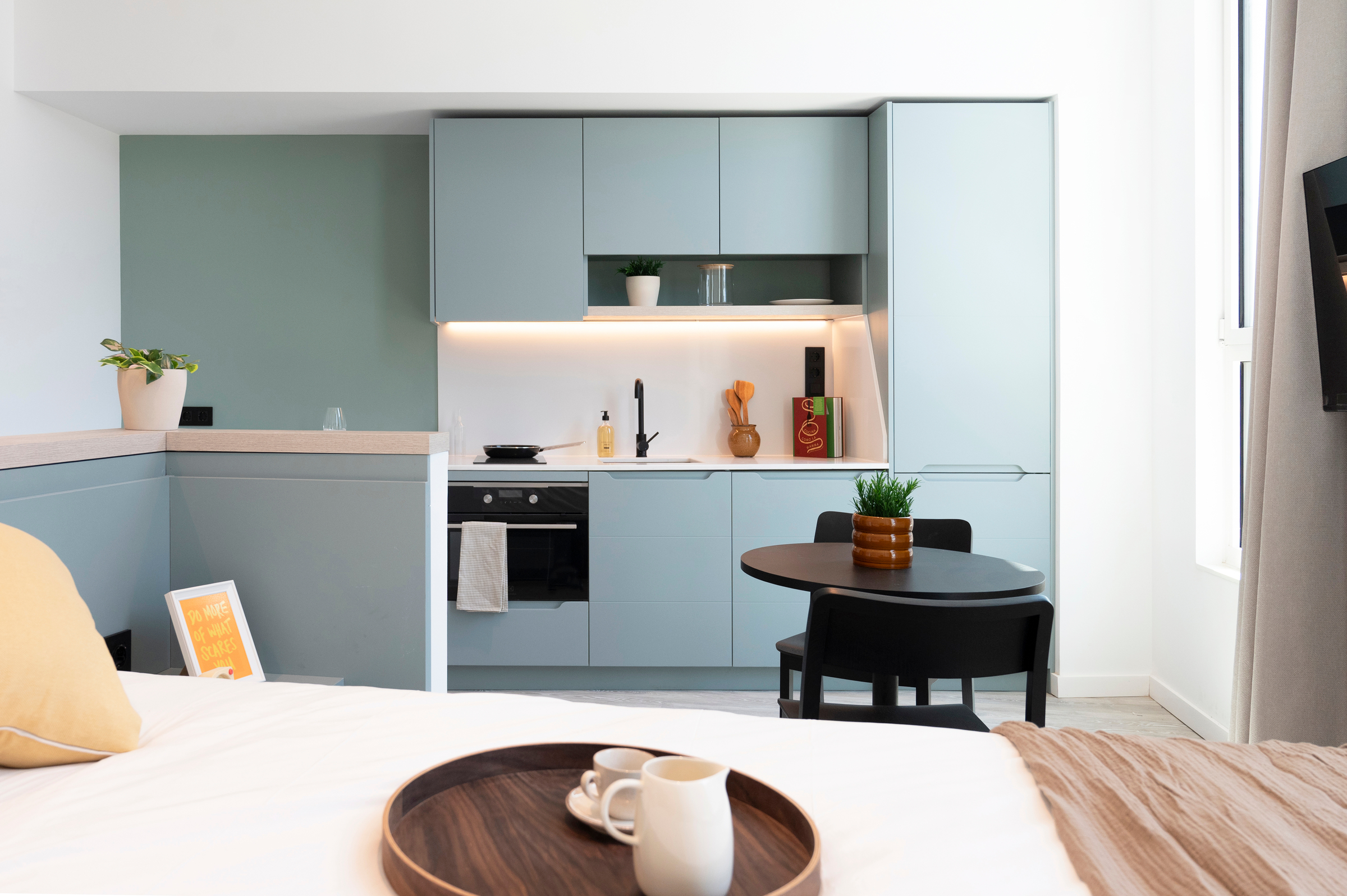 Modern studio apartment with a compact light blue kitchen, round dining table with two chairs, and a neatly made bed in the foreground. The space is bright and minimalistic with plants and soft lighting.