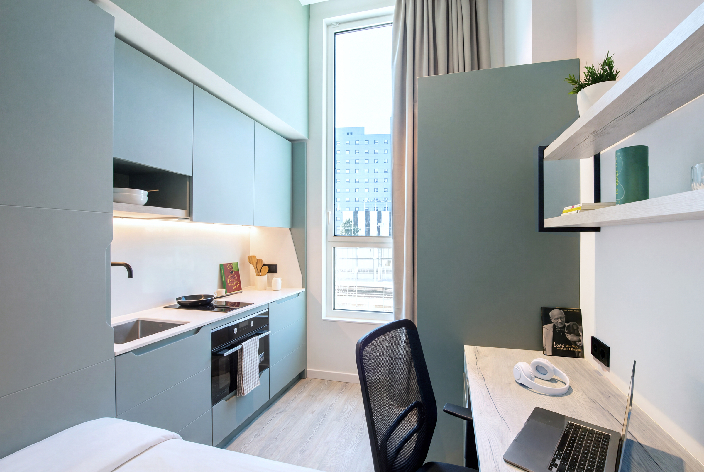 Modern studio apartment with a small kitchen, desk, chair, and large window. Laptop, headphones, and a book are on the desk. Light blue cabinets and minimalist decor create a clean, bright atmosphere.
