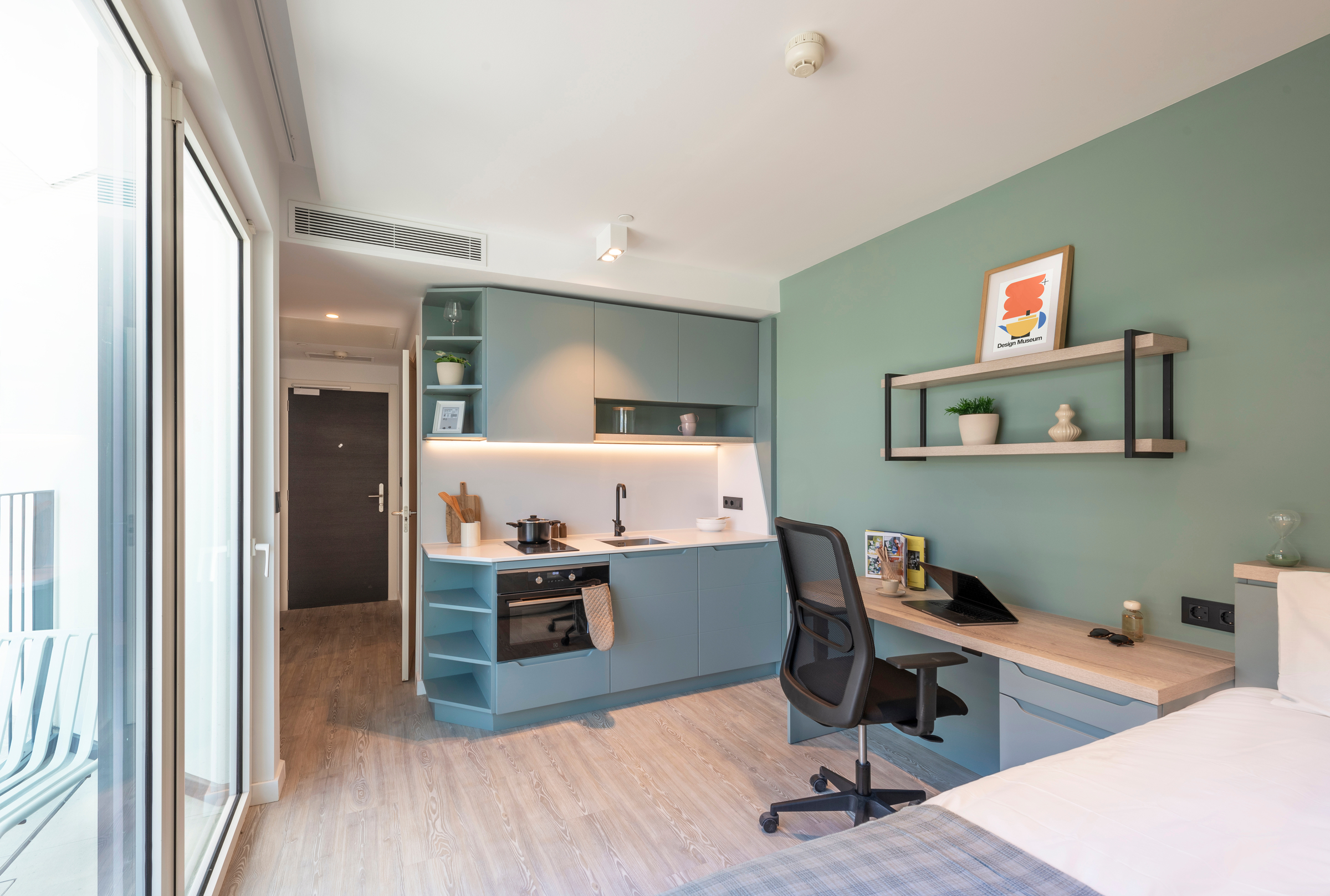 Modern studio apartment with teal cabinets, a small kitchen, desk workspace, shelves, and a bed. Large window provides natural light. Decor includes framed art, plants, and tidy surfaces, creating a cozy and organized atmosphere.