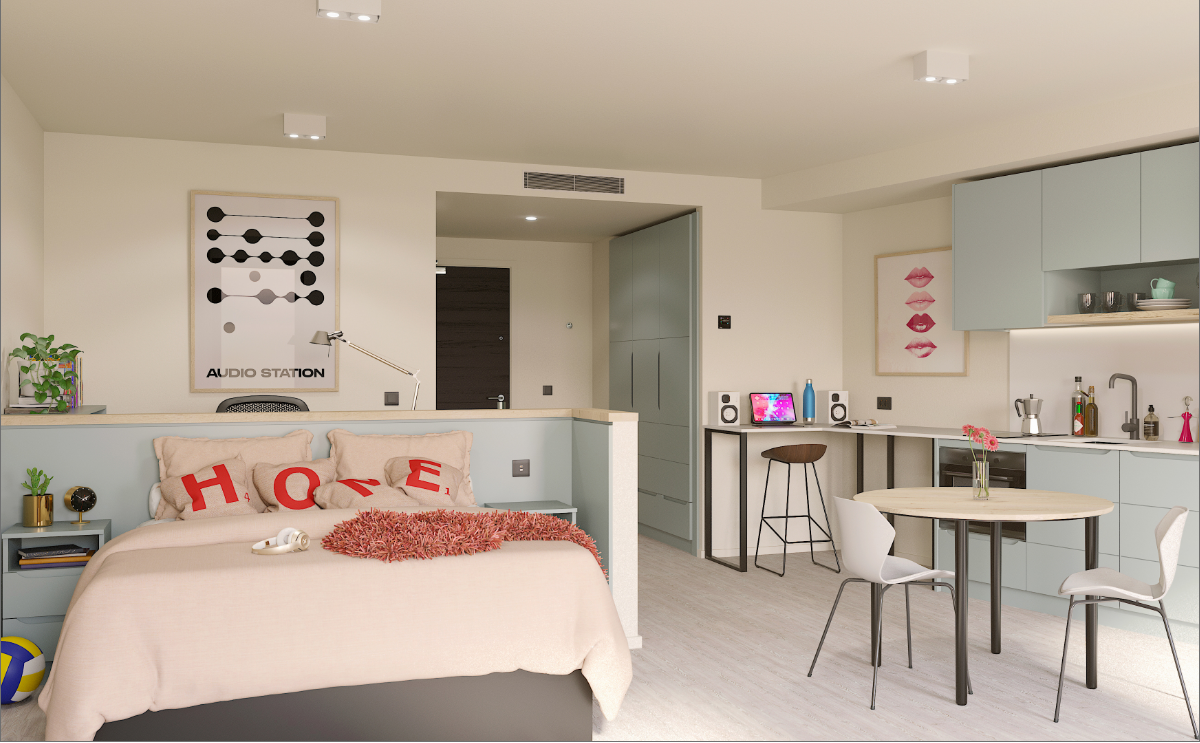 A modern studio apartment with a neatly made bed, decorative pillows spelling "HOME," a small kitchen area, round dining table with two chairs, and wall art. The space is bright with neutral colors and tasteful decor.