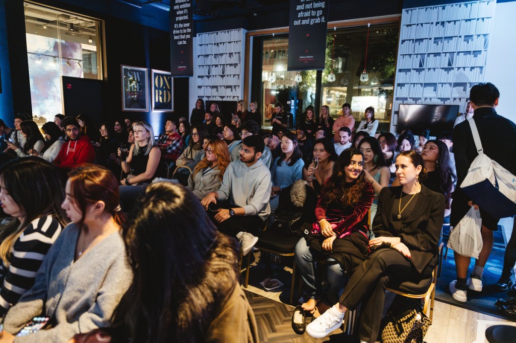 A diverse group of people, possibly students moving to Manchester, sit closely together in rows at an indoor event. Bookshelves and posters decorate the background, while large windows reveal a lit area outside.