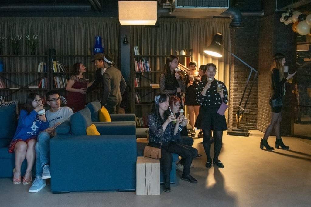 A group of people at a social gathering in cozy, dimly lit Fountainbridge student accommodation, with sofas, bookshelves, and a floor lamp. Some are sitting and chatting; others are standing and holding drinks—an inviting place to live.