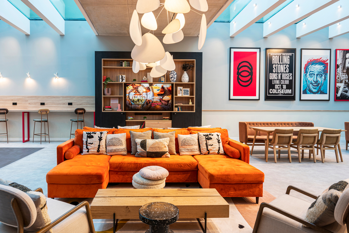 A modern lounge with an orange sectional sofa, wooden coffee table, pendant lights, and bar stools—this space is a great choice for a student looking to decorate a studio apartment with high ceilings, wall art, and bright, colorful decor.