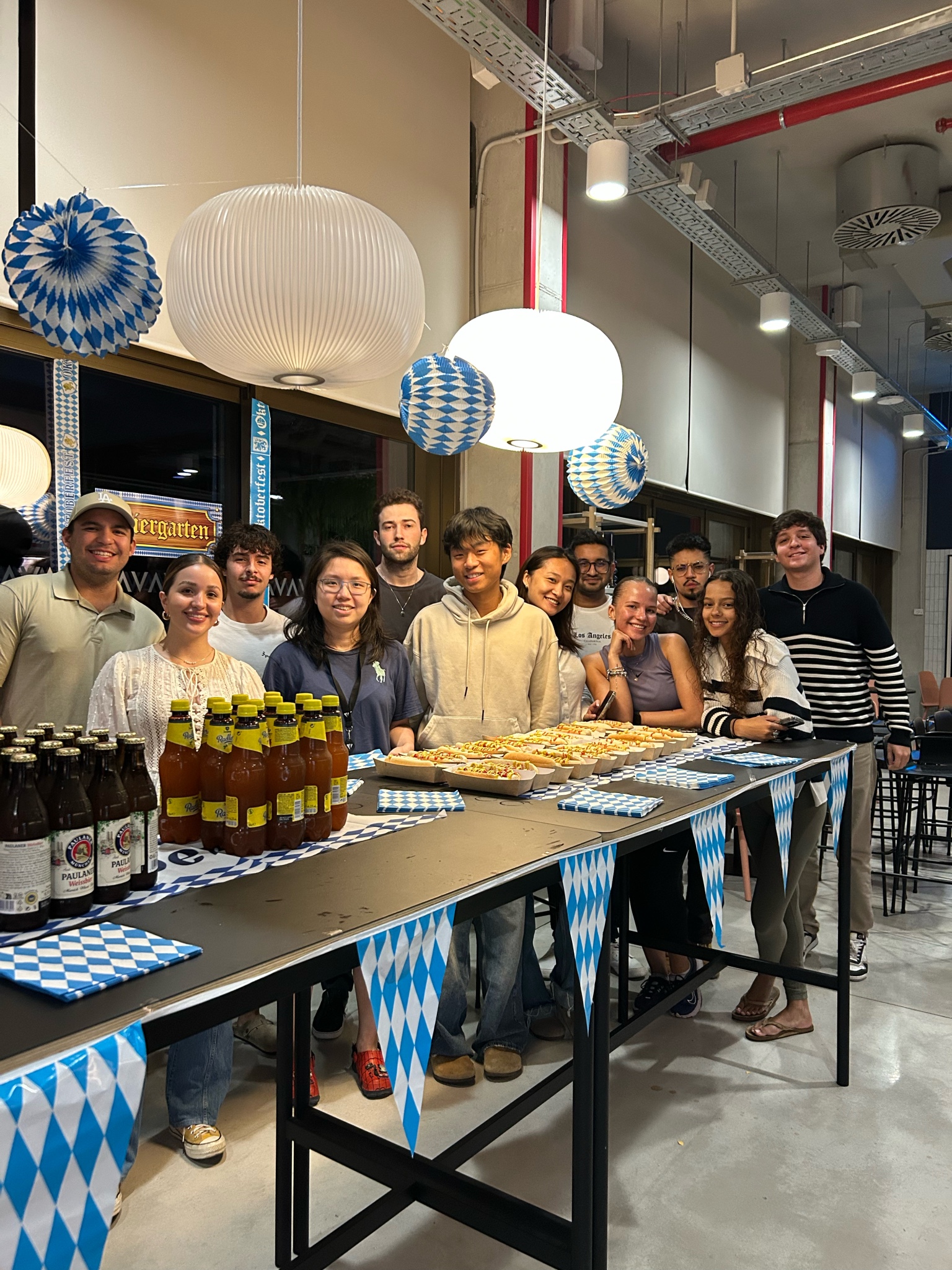 A group of estudiantes internacionales stand smiling behind a table with bottles of beer and trays of pretzels, decorated with blue and white Bavarian patterns, under large round lights in a festively decorated indoor space.