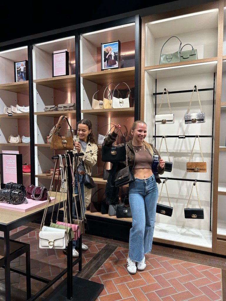 Two young women smile while shopping in a modern, well-lit store, each holding up designer handbags. Behind them, shelves display various purses in different styles and colors—capturing a moment of vida estudiantil stylishly lived.