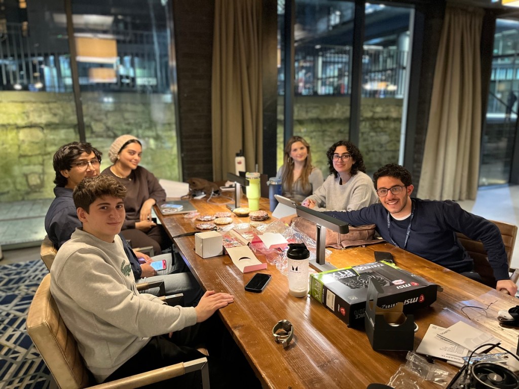 Six young adults, likely students, sit around a wooden table with laptops, snacks, and drink bottles. The cozy indoor setting with large windows suggests a welcoming space in Fountainbridge—a great place to live and collaborate.