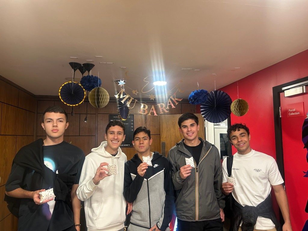 Five young men stand indoors at a Vita Student event, smiling and holding small cards. Behind them, a "EID MUBARAK" banner with blue and gold decorations sets a festive mood, capturing the celebratory spirit of eventos en Vita Student.