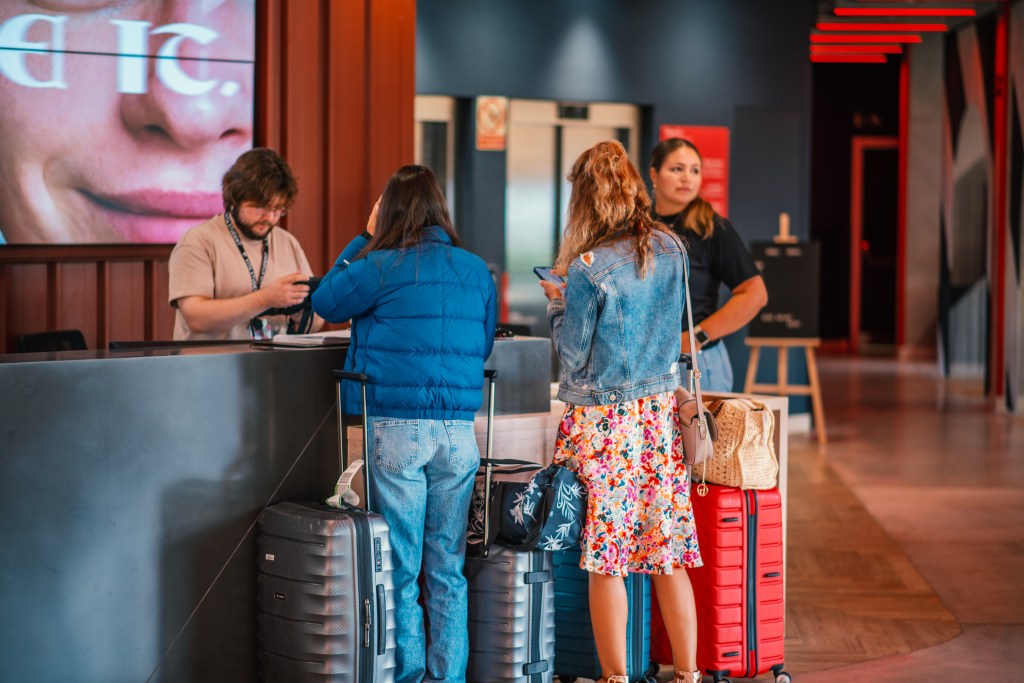 Three women with suitcases stand at a hotel reception desk while a staff member assists them, likely discussing trámites esenciales for estudiantes internacionales. The modern setting is bright, with a large digital screen partially visible in the background.