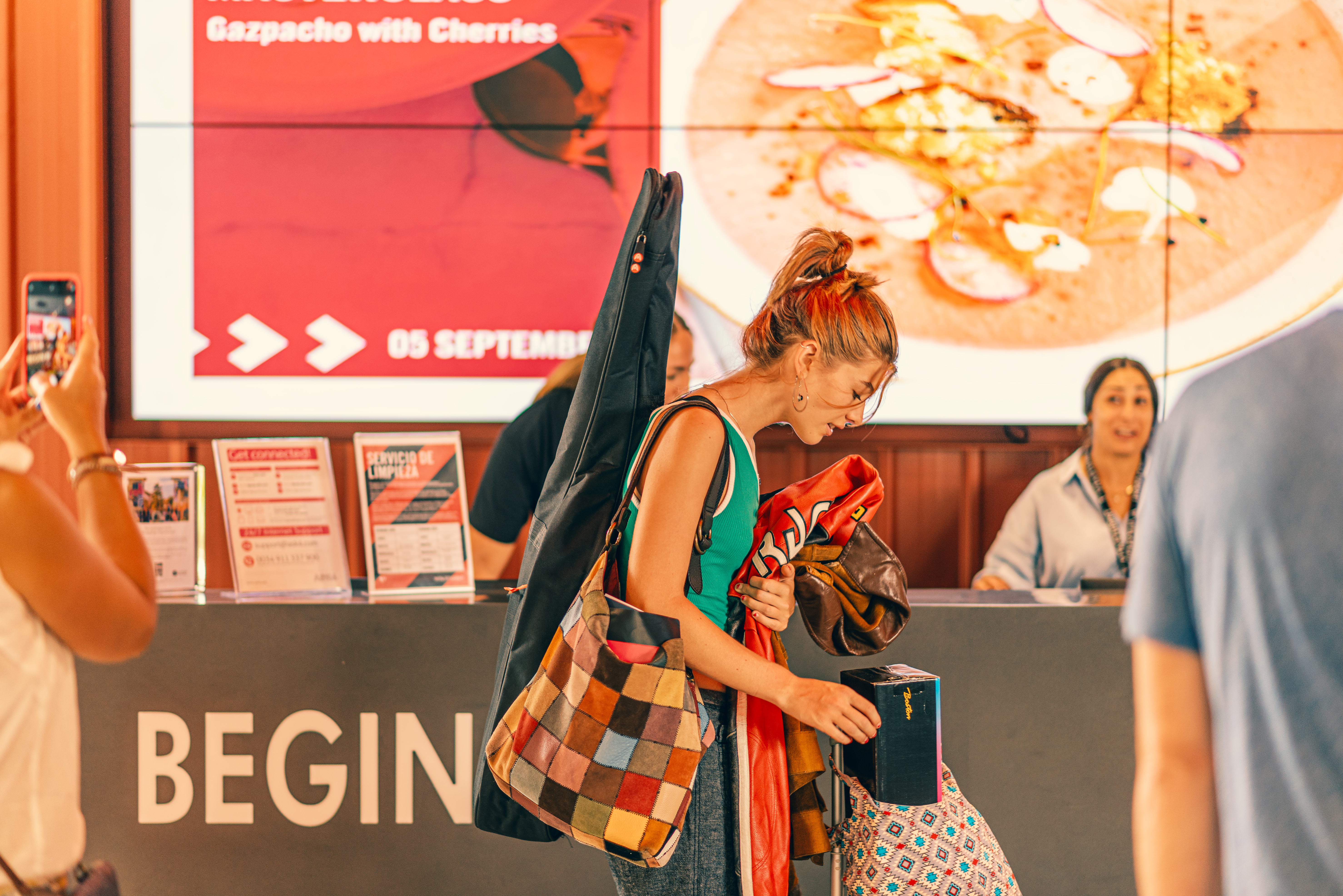 A woman with a guitar case and colorful bags stands in front of a counter, holding a passport—likely handling trámites esenciales for estudiantes internacionales. Behind her, a digital screen shows food and event info; people and staff are visible in the background.