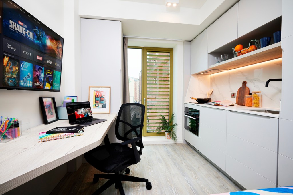 A modern studio apartment features a cozy workspace with a laptop, notepads, and pens—making it a great choice for students. Enjoy a wall-mounted TV with streaming apps, a compact kitchen with white cabinets, and a small plant by the window.