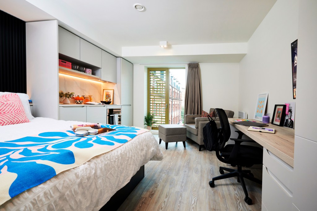 A modern, tidy bedroom in a studio apartment with a bed, desk, chair, shelves, small sofa, and large glass doors opening to a balcony. With light wood flooring and bright natural light, it's a great choice for students.