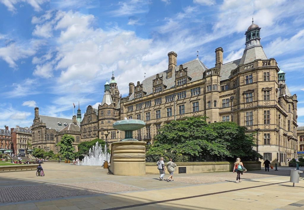 A large historic stone building with turrets and tall windows stands behind a plaza with a fountain, trees, and people enjoying student-friendly things to do under Sheffield’s blue sky with scattered clouds.