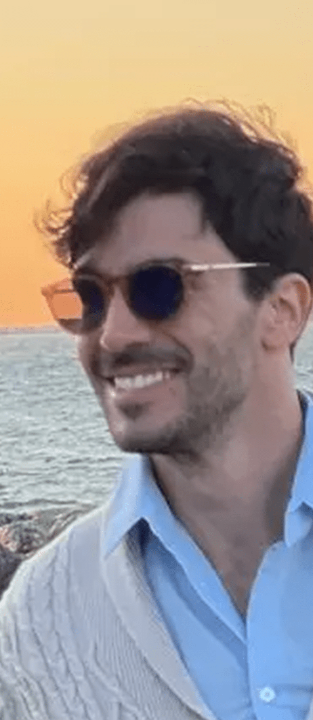 A man with dark hair and sunglasses smiles while standing by the ocean at sunset, wearing a light blue shirt and cream sweater, enjoying a well-deserved break after exploring new career tools.