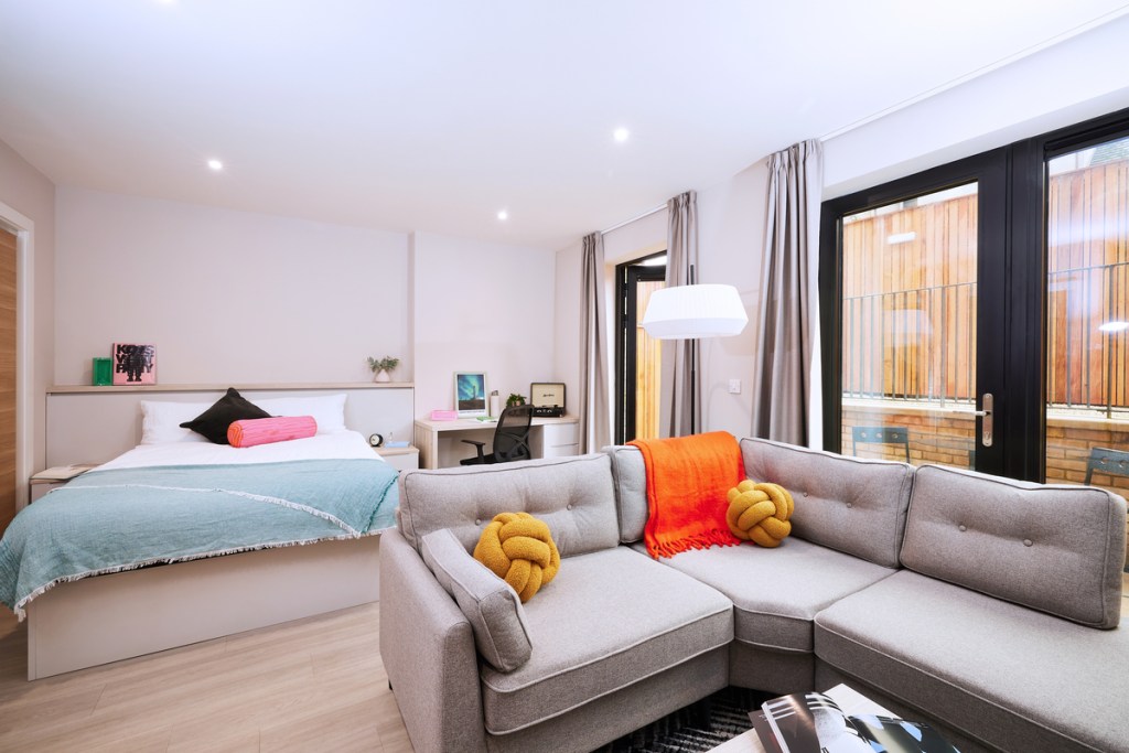 A modern studio apartment with a bed, desk, and gray sectional sofa decorated with yellow knot pillows and an orange throw. Large windows brighten the space—making it a great choice for any student seeking comfort and style.