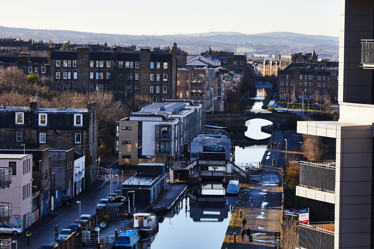 A canal runs through Fountainbridge, a popular place to live for students, with modern and older apartments on either side reflecting the morning sunlight. Pedestrians stroll along the towpath, and bridges span the water, with hills visible in the distance.