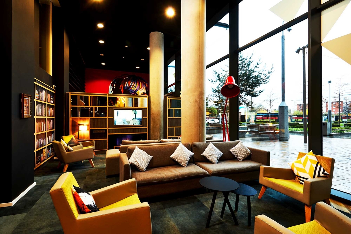 Modern lounge with large windows, grey sofas, yellow armchairs, and a red floor lamp—an inviting space for students. A bookshelf and decorative pillows add charm. Warm lighting makes this a great choice for any studio apartment. Trees and streetlights are visible outside.