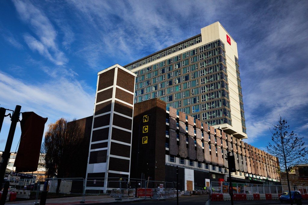 A multistory building complex with NCP parking signs, featuring a tall modern tower behind a shorter brick structure—possibly Vita Student Sheffield—under blue skies. Construction barriers and fencing are visible in the foreground.