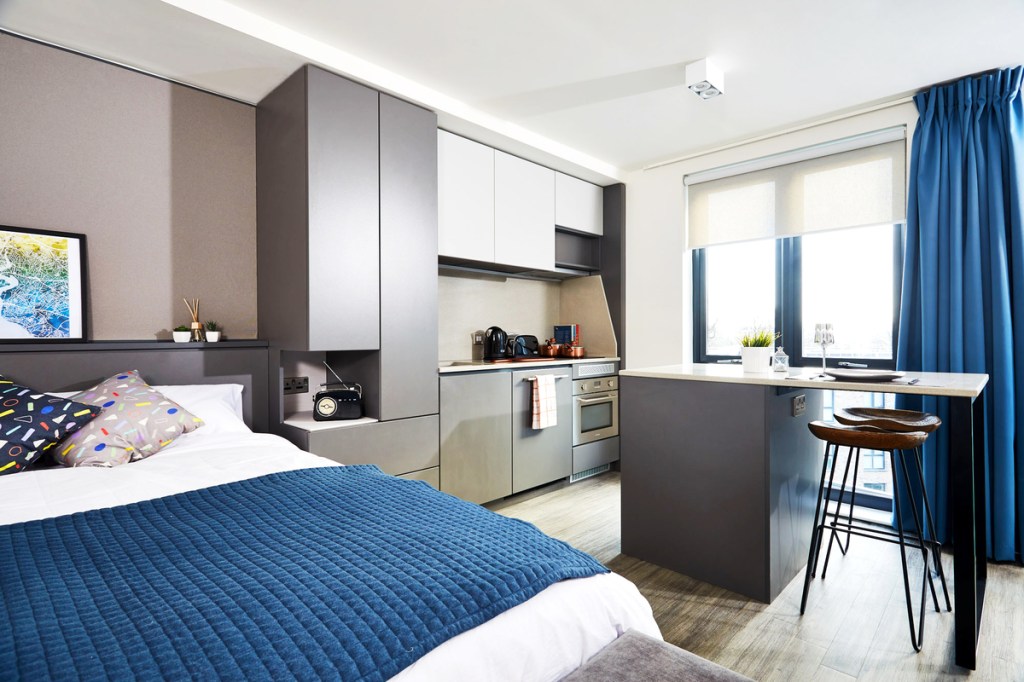 Modern studio apartment with a bed, blue quilt, cozy pillows, compact kitchen, bar-style dining area, and large windows with blue curtains. A great choice for any student seeking comfort and convenience in one stylish space.