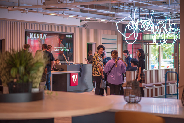 A group of people stand and chat near a reception desk in a modern, brightly lit Vita Student office lobby with neon lights and contemporary decor. A digital screen displays the day and motivational event details.