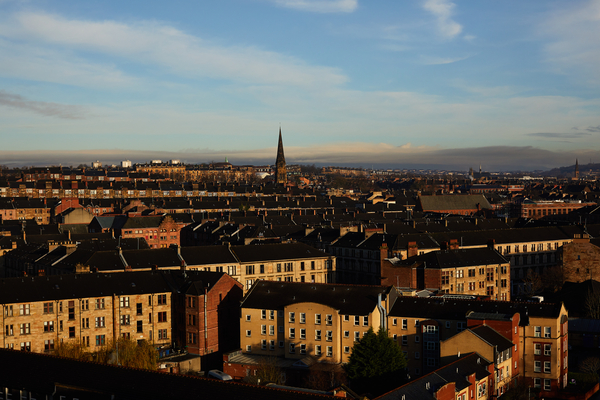 A cityscape with rows of tan and brown buildings under a blue sky, featuring a church with a tall, pointed steeple—capturing the essence of student life and the unique charm of living in Glasgow, all bathed in warm late afternoon sunlight.