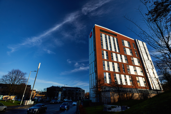 A tall, modern building with red and white panels stands beside a busy street in Glasgow, surrounded by leafless trees and smaller buildings—a familiar scene for those experiencing student life under the city’s clear blue sky.