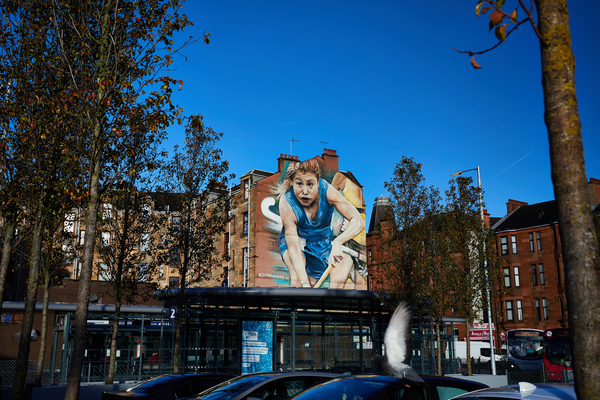 A large mural of a female athlete holding a hockey stick is painted on the side of a building, highlighting Glasgow student life in an urban area with trees, parked cars, and a bus stop in the foreground.