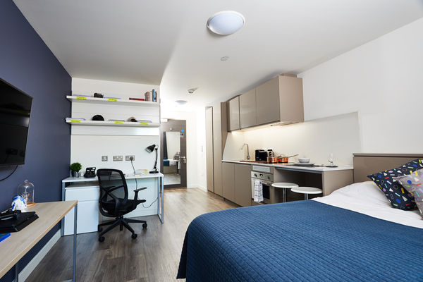 Enjoy modern student life in this bright Glasgow studio apartment, featuring a blue bedspread, kitchenette, two stools, desk with chair, shelves, and a TV on a blue accent wall—all designed for tidy, minimalist living.