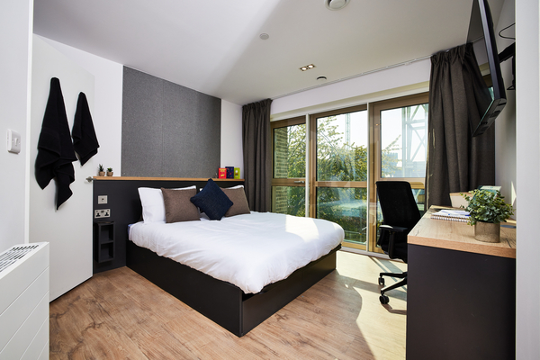 Modern bedroom with a large bed, desk and chair by the window, wooden floor, dark curtains, wall-mounted TV, and a small plant on the desk—offering a glimpse into student life London with natural light pouring in through glass doors.